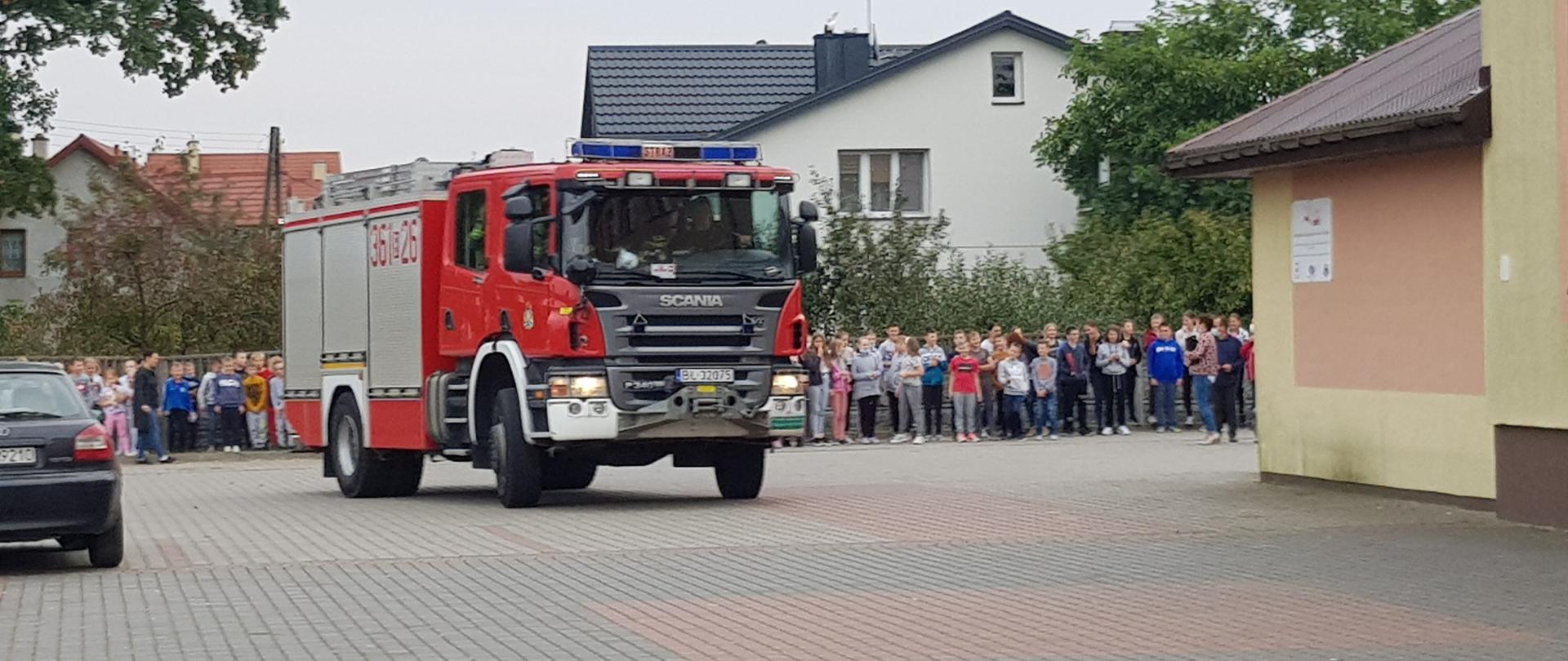 Na pierwszym planie samochód gaśniczy dojeżdżający do szkoły. W tle ewakuowana młodzież stojąca na placu szkoły