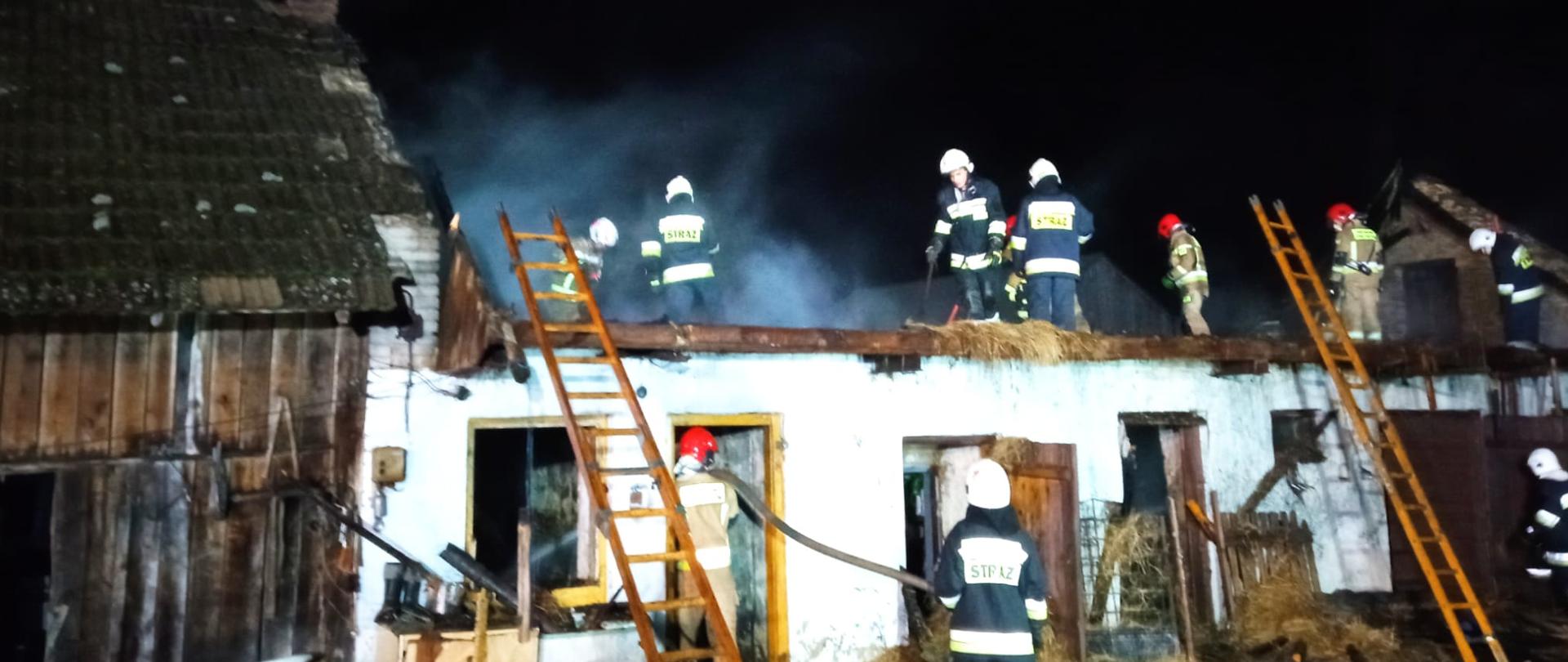 Zdjęcie przedstawia strażaków na dachu budynku gospodarczego - jest noc, w tle panuje zadymienie. Teren akcji jest mocno oświetlony. Do budynku przystawione są drabiny. Przed budynkiem stoją strażacy - trzymają linie gaśnicze. Przed budynkiem widać porozrzucane siano.