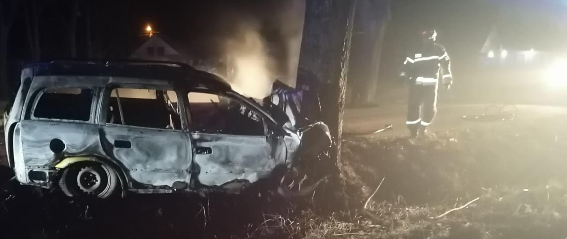 Przy drzewie stoi cały spalony samochód typu combi z rozbitym przodem. w oddali widać strażaka oraz budynek mieszkalny. 