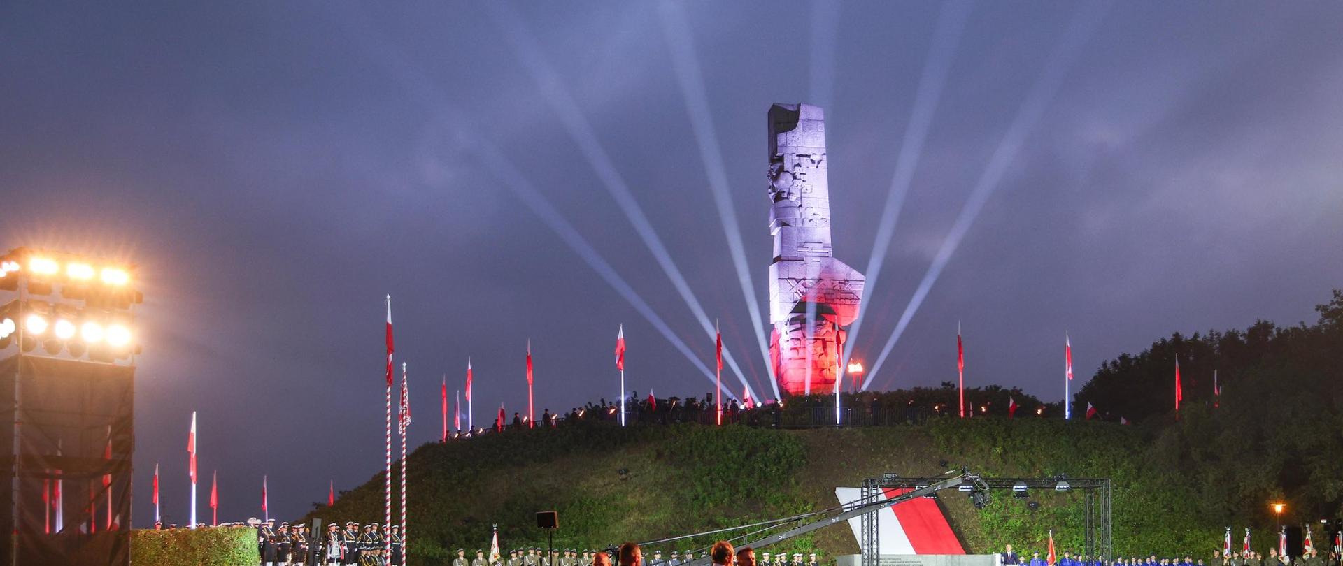 Uroczystości pod Pomnikiem Obrońców Wybrzeża na Westerplatte w Gdańsku. Monument jest podświetlony w barwach biało-czerwonych, a w niebo skierowane są jasne snopy światła. W pierwszym planie zgromadzeni są wojskowi i goście, a dalej stoją poczty sztandarowe. Wokół widoczne są polskie flagi.
