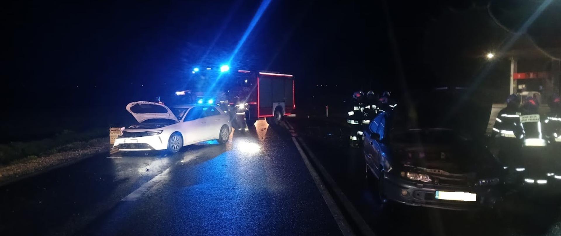 Na zdjęciu widać w porze nocnej dwa osobowe auta rozbite na jezdni. W tle samochód strażacki.