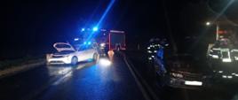 Na zdjęciu widać w porze nocnej dwa osobowe auta rozbite na jezdni. W tle samochód strażacki.