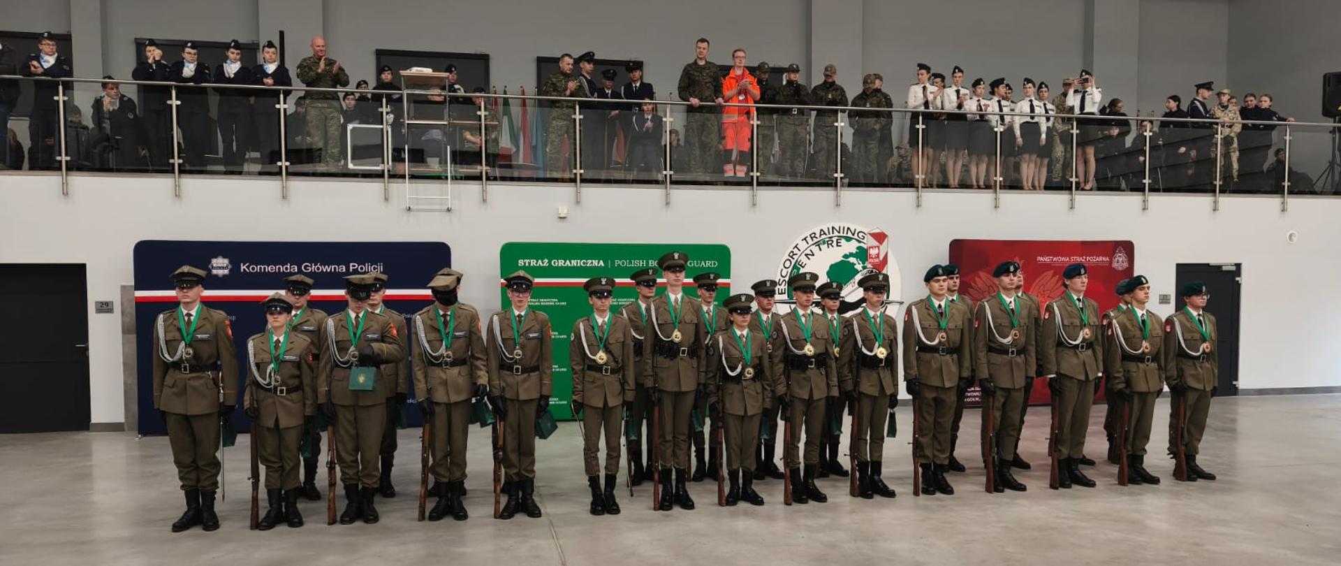 Finaliści stoją w szeregu frontowym z medalami; za nimi balkon z publicznością i ścianki Komendy Głównej Policji, Straży Granicznej i PSP.