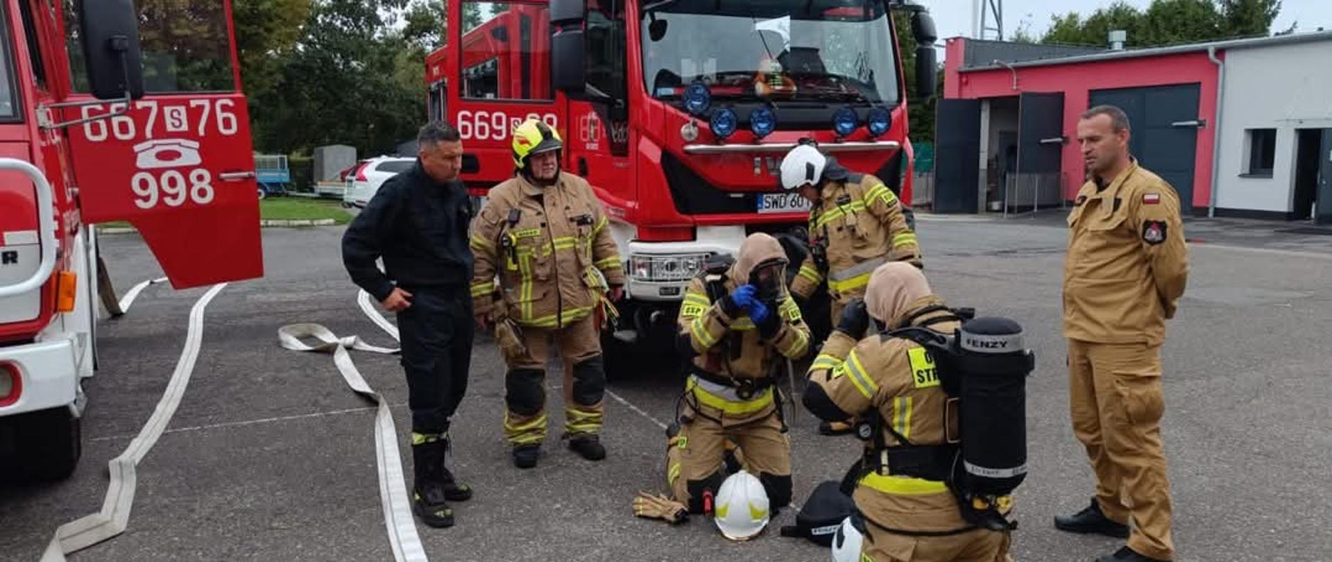 Strażacy ubierają się w aparaty powietrzne podczas przygotowania do ćwiczeń na tle samochodów pożarniczych