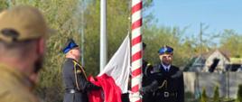 Trzech strażaków w galowych mundurach stoi przy czerwono-białym maszcie flagowym. Dwóch z nich trzyma i rozwija biało-czerwoną flagę, przygotowując ją do podniesienia, a trzeci stoi obok na baczność. Na pierwszym planie, lekko rozmyty, widoczny jest mężczyzna w brązowym stroju z naszywką Polski na ramieniu, obserwujący ceremonię. W tle znajdują się drzewa, trawa oraz zabudowania, a na dole widnieje emblemat Państwowej Straży Pożarnej z napisem „Gryfice”.