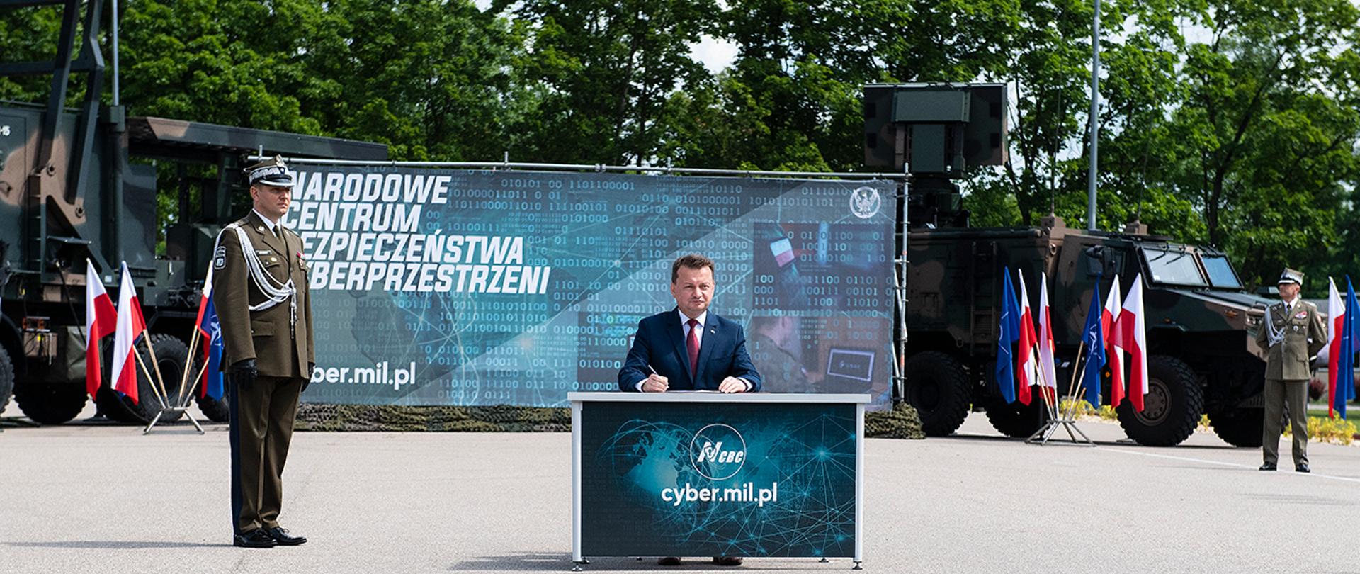 Szef MON podpisuje dokument na placu apelowym NCBC. W tle baner, żołnierze i sprzęt wojskowy.