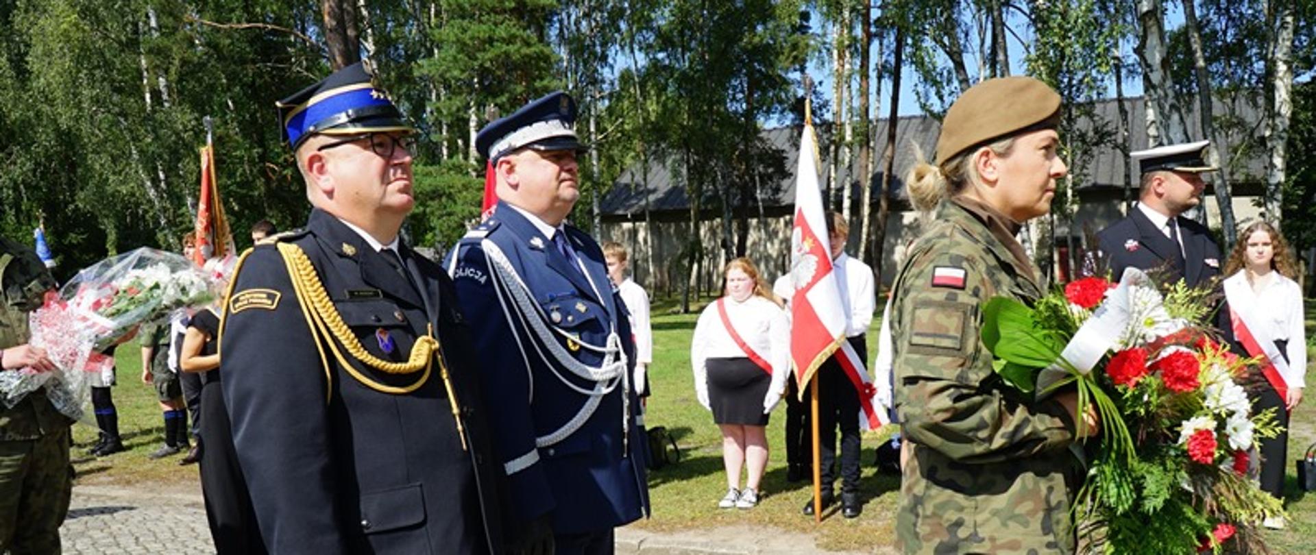 Żołnierz niesie wiązankę kwiatów za nim stoją przedstawiciele Policji oraz Państwowej Straży Pożarnej obok stoją poczty sztandarowe.