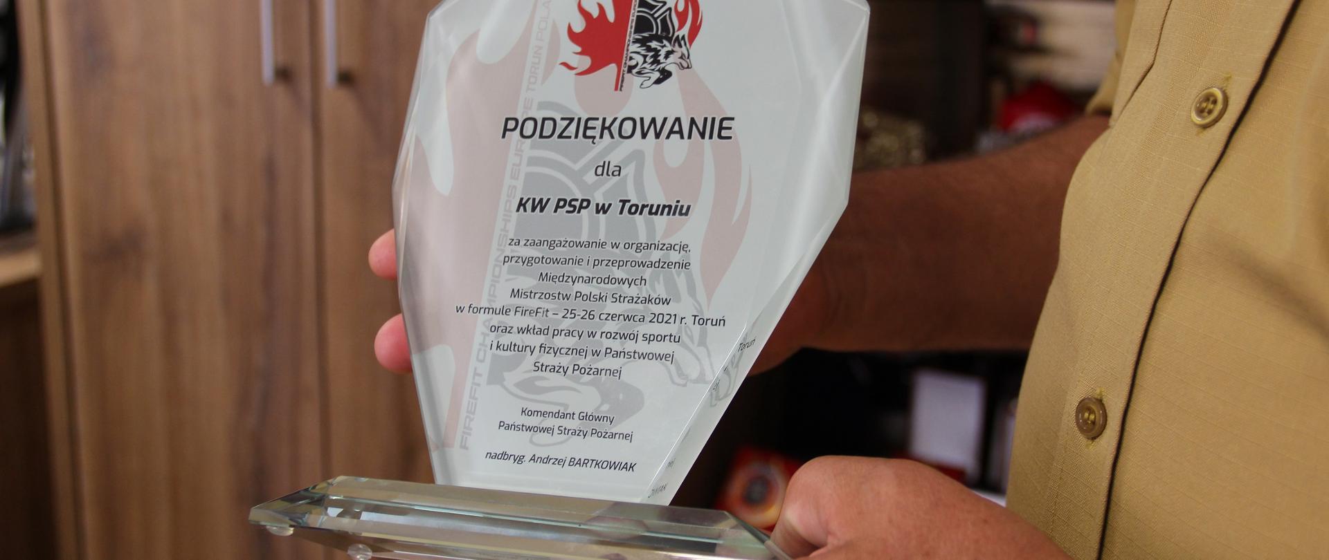 Na dłoni komendanta wojewódzkiego statuetlka z dedykacją i logo mistrzostw. W tle elementy wyposażenia gabinetu.