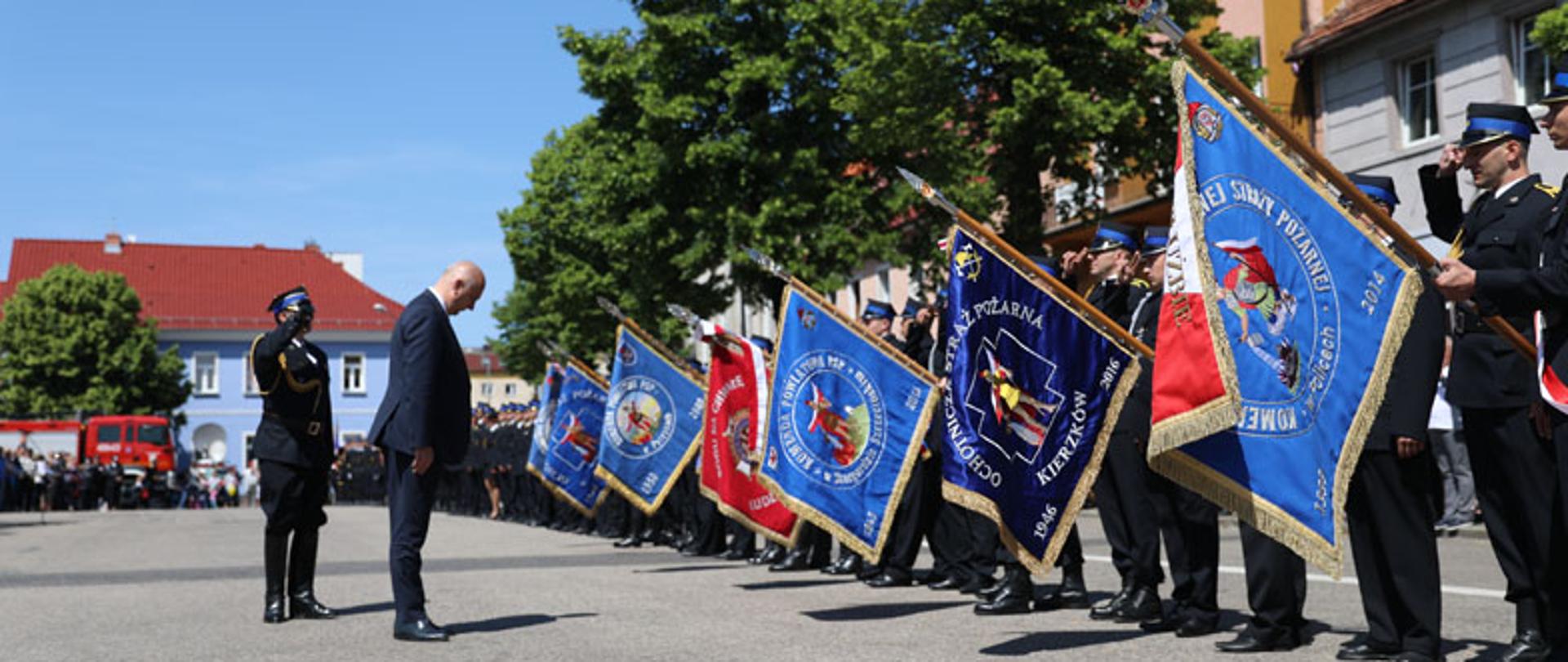 Człowiek z pochyloną głową przed szpalerem strażaków ze sztandarami
