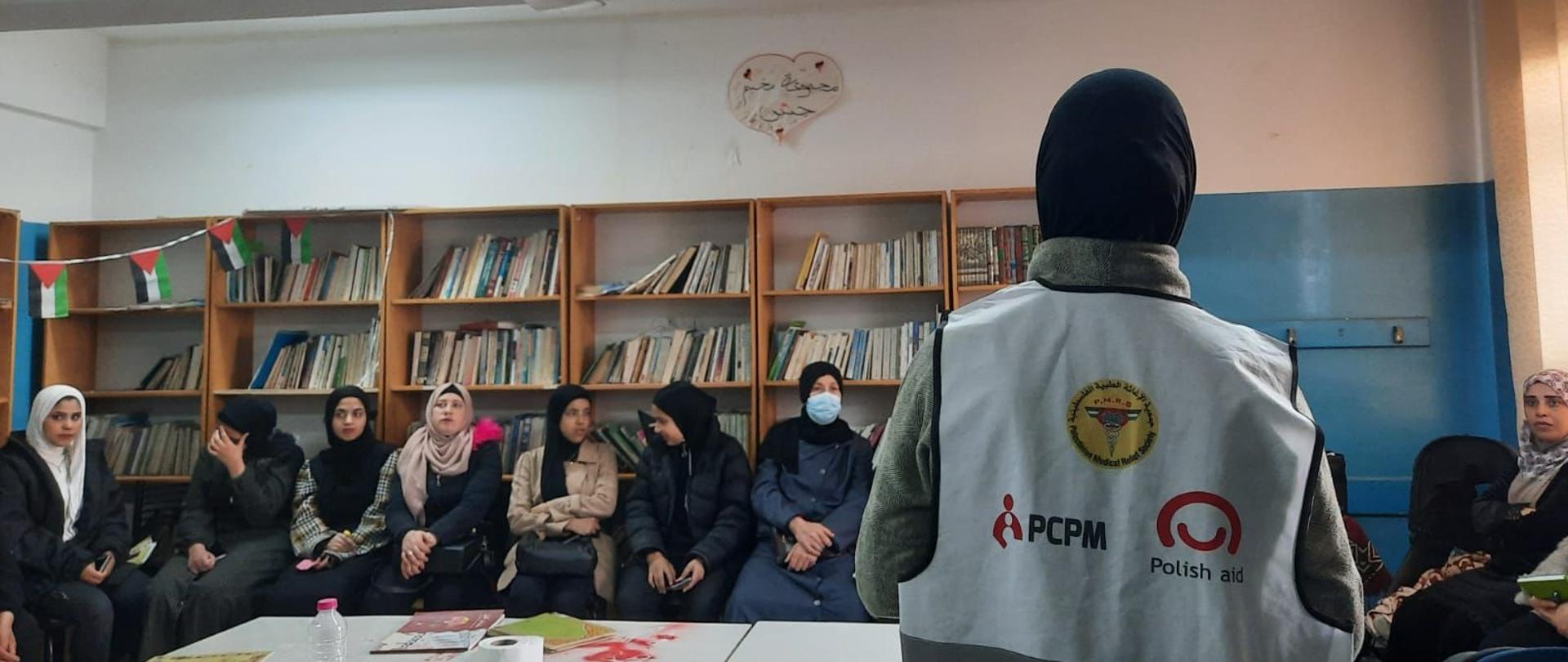 W sali siedzą kobiety w hidżabach i słuchają instruktorki prowadzącej szkolenie