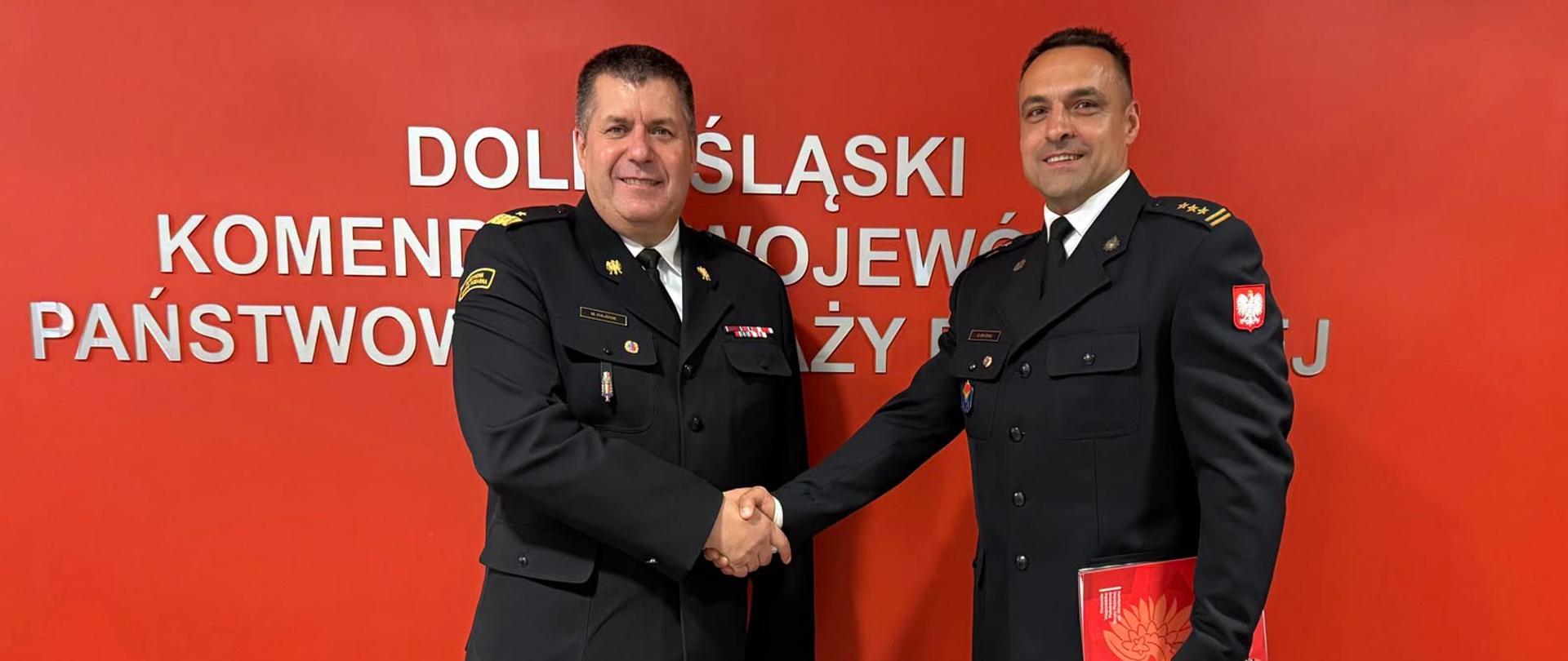 Strażacy w ciemnych mundurach na tle jaskrawoczerwonej ściany z białym napisem "Dolnośląski Komendant Wojewódzki Państwowej Straży Pożarnej"