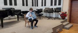 Chłopiec siedzi na krześle i gra na akordeonie, na podwyższonej scenie, w tle fortepian, krzesła i pulpity.