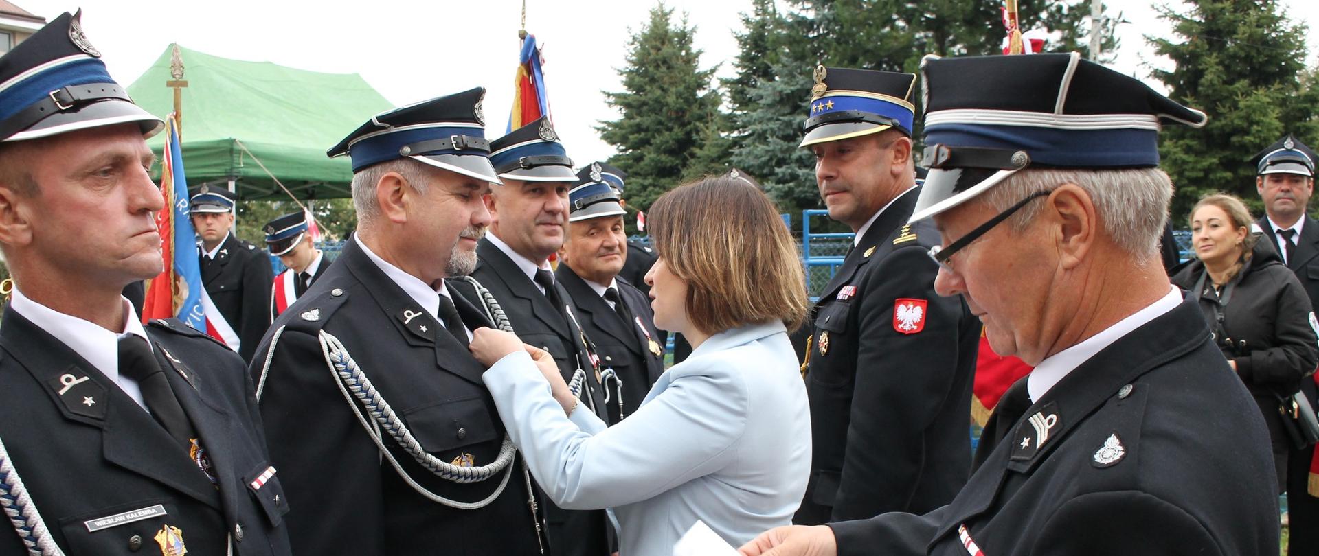Zdjęcie przedstawia moment wręczenia medali druhom OSP przez Poseł na Sejm Annę Schmidt. W tle widoczni są zaproszeni goście, druhowie oraz poczty sztandarowe. Strażacy ubrani są w umundurowanie wyjściowe ze sznurem.