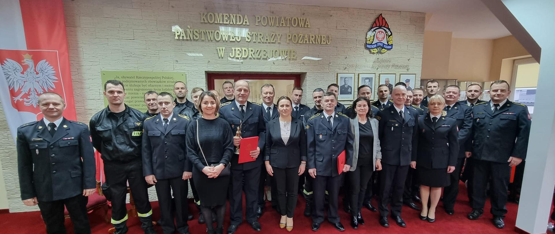 Funkcjonariusze i pracownicy Komendy Powiatowej Państwowej Straży Pożarnej w Jędrzejowie na uroczystej zbiórce pożegnania emerytów. Wszyscy znajdują się na sali tradycji.