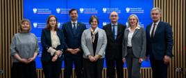 Zdjęcie zbiorowe, pod ścianą z pionowych drewnianych listewek z wbudowanym wielkim ekranem stoi siedem osób, pośrodku wiceminister Zioło-Pużuk.