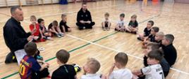 Pogadanka młodych zawodników ze strażakiem na temat zagrożeń mogące wystąpić zarówno w domu, jak i w codziennym życiu. Dzieci siedzą na parkiecie hali sportowej w kółku wysłuchując porad strażaka.