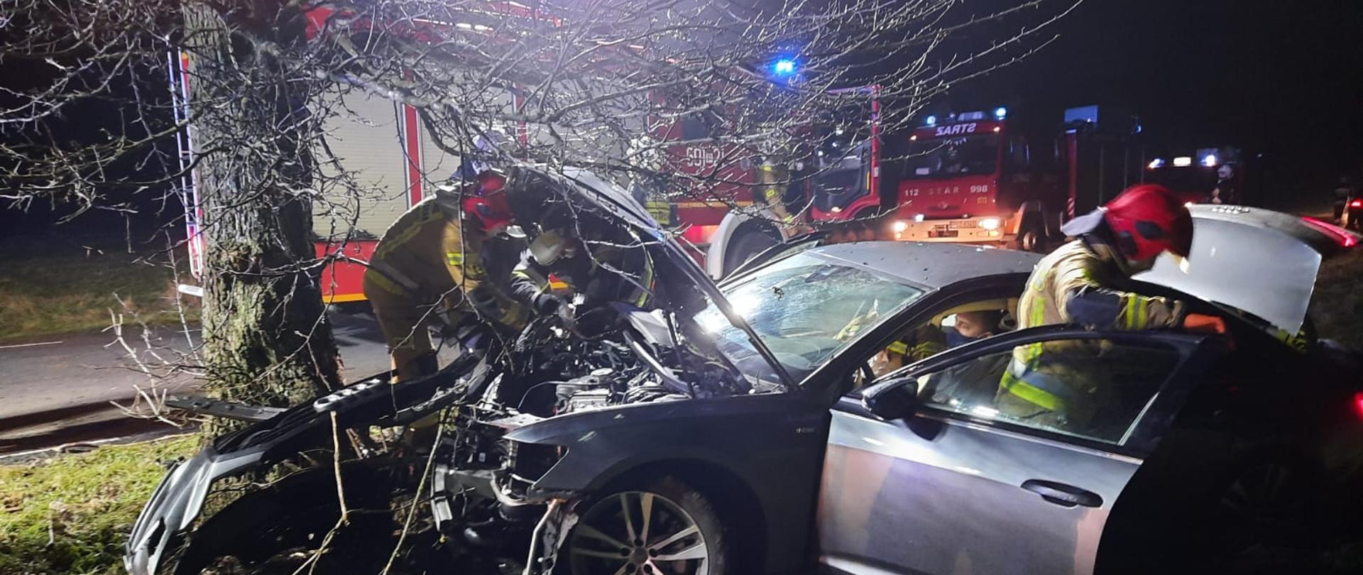 Widać samochód z uszkodzonym przodem, który jest blisko drzewa, za nim są samochody straży