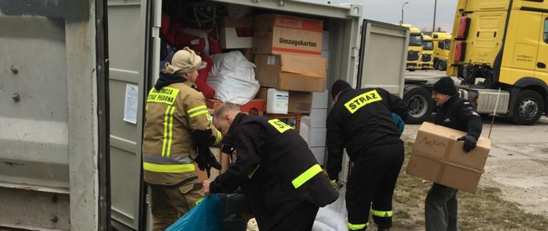 Na zdjęciu biały kontener morski. Wewnątrz kartony i różne opakowania. Przed strażacy w czarnym umundurowaniu i osoby postronne. Po prawej stronie żółty ciągnik siodłowy. Zachmurzenie duże.