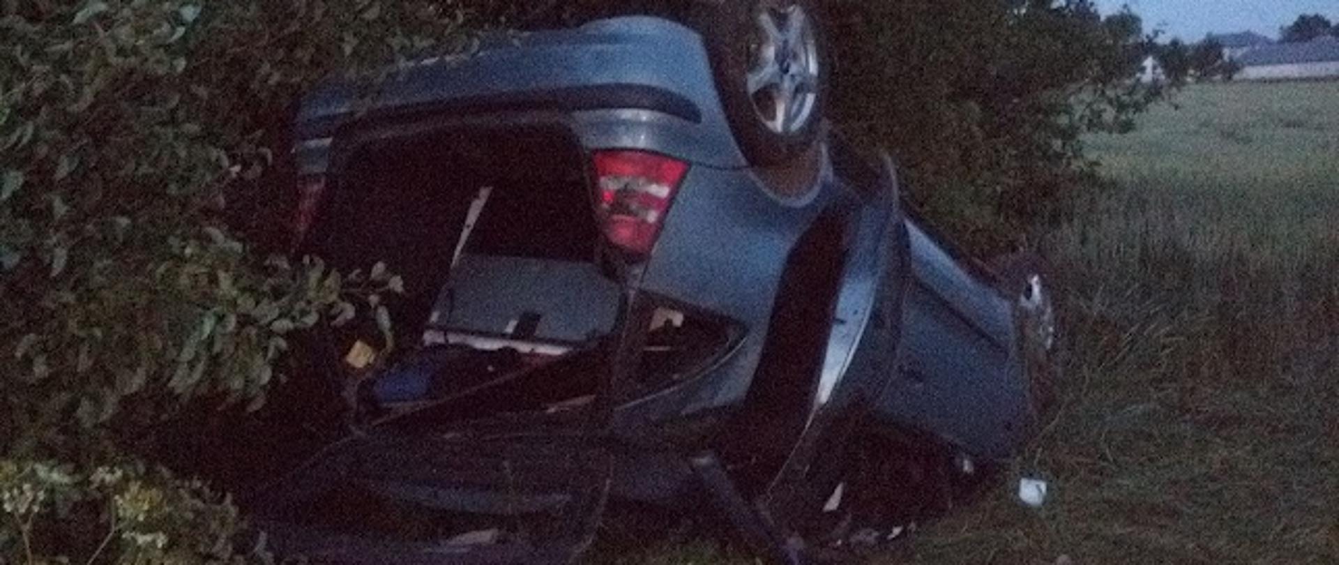 Leżący na poboczu jezdni na dachu samochód osobowy. Wokół porozrzucane elementy wyposażenia i karoserii.