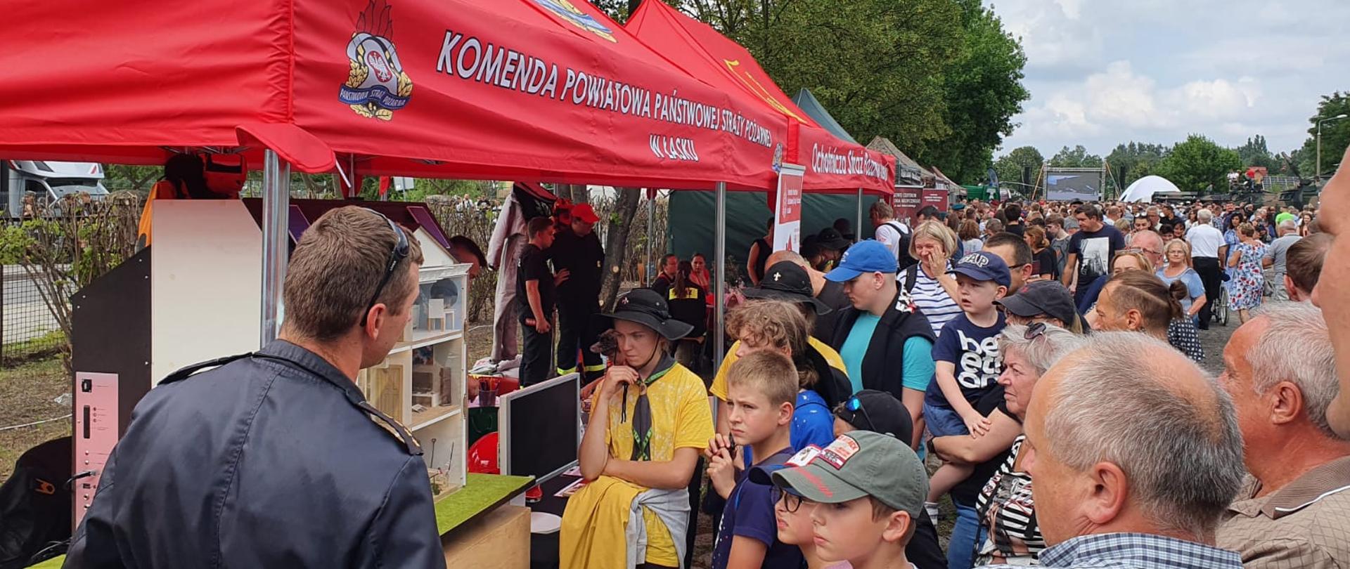 Zdjęcie przedstawia strażaka podczas prezentacji stoiska na pikniku wojskowym zorganizowanym z okazji święta Wojska Polskiego, upamiętniającego Cud nad Wisłą. 