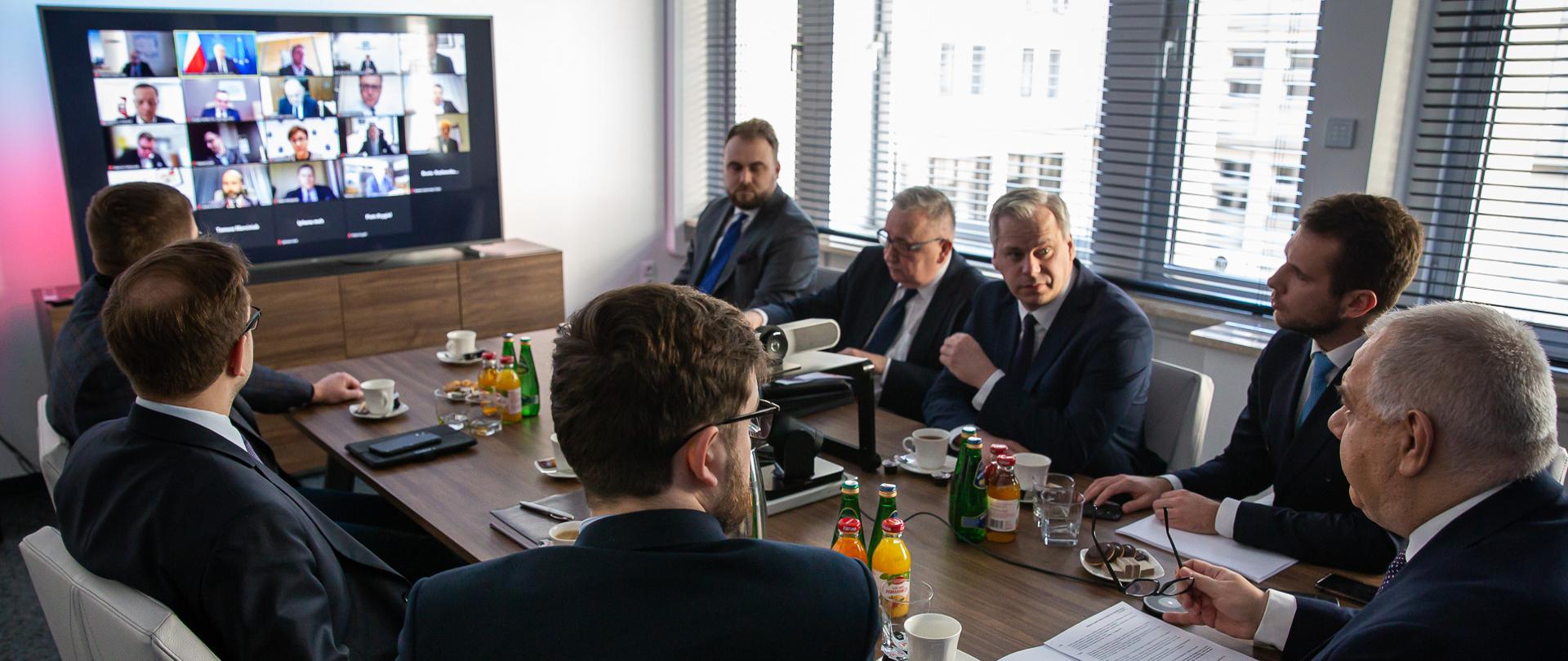 Przy stole siedzi wicepremier Jacek Sasin oraz wiceministrowie: Jan Kanthak, Karol Rabenda, Piotr Pyzik, Andrzej Śliwka, Maciej Małecki oraz szef zespołu Synergia Łukasz Turkowski i dyrektor Biura Ministra Piotr Rycerski. W tle ekran z okienkami łączenia z przedstawicielami spółek.