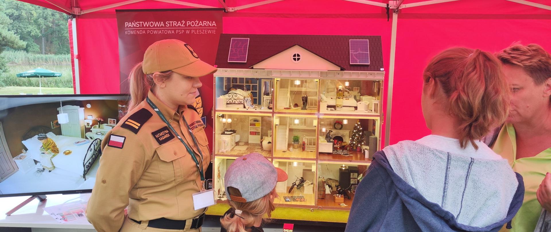 Kobieta ubrana w białą kuszulę i strażacką rogatywkę opowiada dwójce dzieci i dwójce dorosłych o zagrożeniach znajdujących się w mobilnym symulatorze zagrożeń