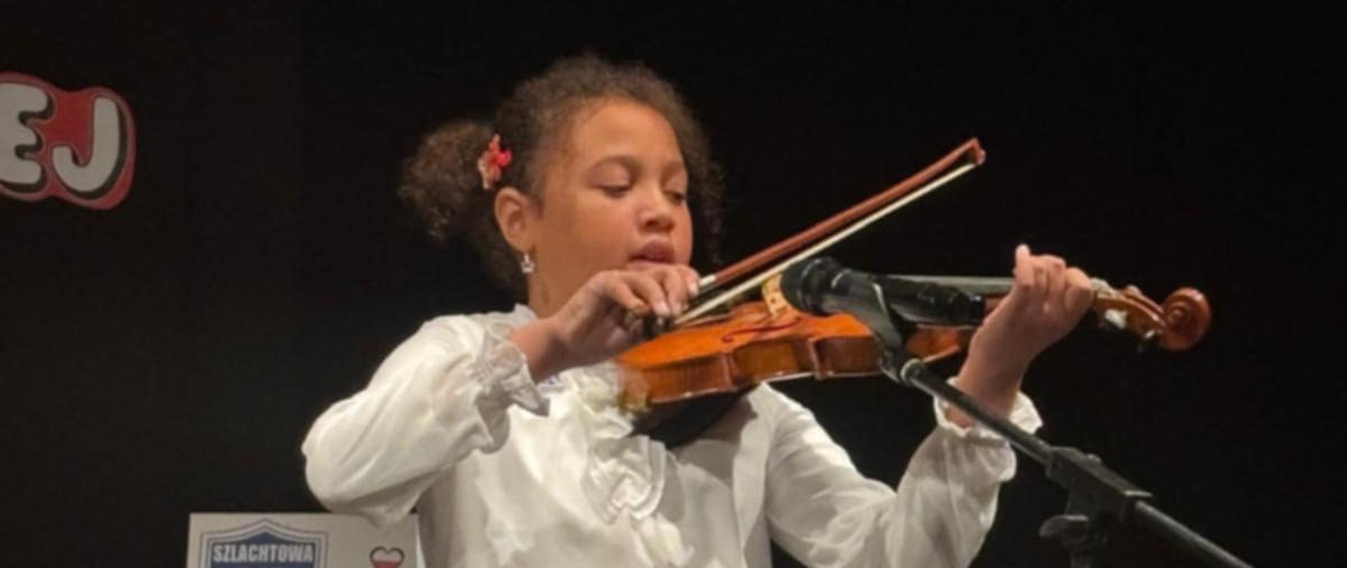 Na zdjęciu widać młodą dziewczynkę grającą na skrzypcach na scenie. Stoi prosto, skupiona na grze, trzyma skrzypce pod brodą i porusza smyczkiem po strunach. Ma ciemne, kręcone włosy związane w dwa kucyki, a w jednym z nich widoczna jest czerwona spinka w kształcie kwiatu. Ubrana jest elegancko — ma białą koszulę z długim rękawem i czarną, plisowaną spódniczkę. Na nogach ma czarne rajstopy. Przed nią stoi mikrofon, a w tle, po lewej stronie, znajduje się plansza z rysunkiem dziewczynki w biało-czerwonym stroju i napisem „Szlachtowa SP”. Scena jest ciemna, oświetlona światłem skupionym na wykonawczyni, co podkreśla, że to występ lub konkurs muzyczny.