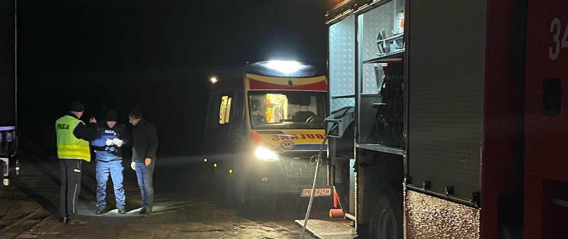 Na zdjęciu widzimy bok samochodu strażackiego koloru czerwonego, otwarte srebrne żaluzje, Do pojazdu podłączony jest wąż koloru czarnego. Dalej w tle widzimy ambulans koloru żółtego.