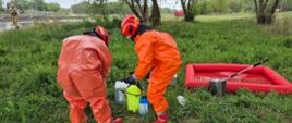 Dwóch Strażaków w kombinezonach ochronnych pochyla się nad sprzętem do prowadzenia badań. Stoją na trawie, niedaleko brzegu rzeki.