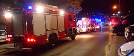 Na zdjęciu widać trzy samochody strażackie stojące na ulicy przed budynkiem z włączonymi niebieskimi sygnałami. Przy jednym pojeździe widać grupę strażaków, którzy przebierają się po akcji.