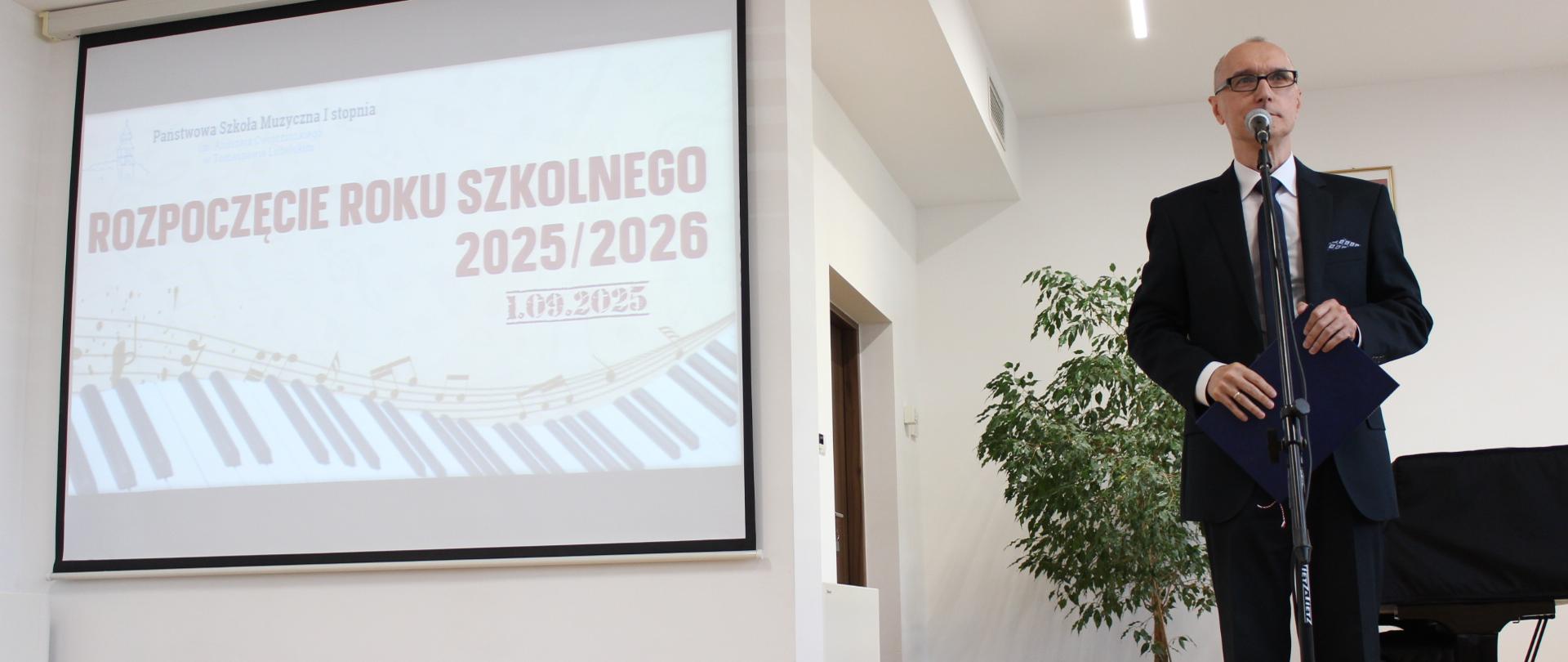 na estradzie po lewej projektor z wyświetlanym plakatem na zakończenie roku szkolnego, z prawej strony dyrektor przemawiający do mikrofonu