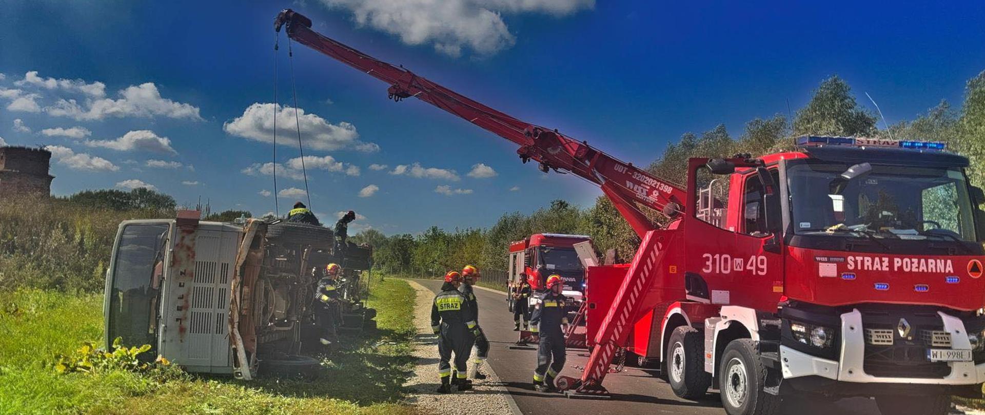 Na zdjęciu czerwony samochód strażacki z dźwigiem podnoszący przewrócony samochód ciężarowy, leżący na boku. Między pojazdami grupa strażaków w ciemnych mundurach i czerwonych hełmach. Nad terenem akcji widoczne jest pogodne błękitne niebo z białymi chmurami. 