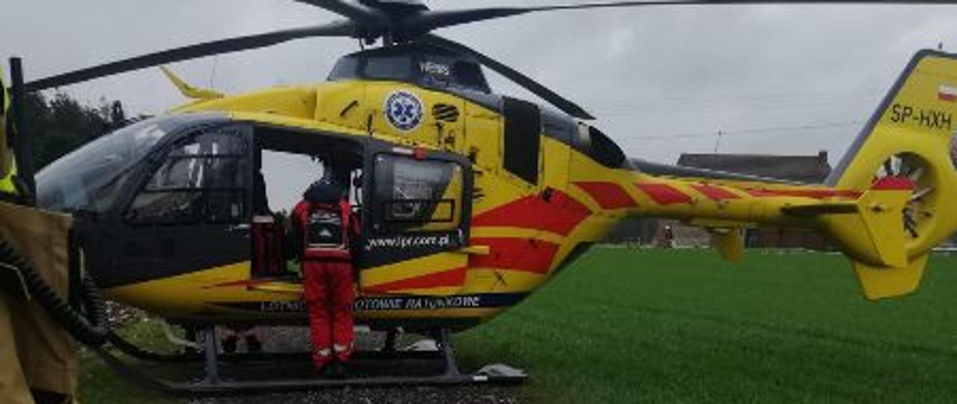 Na zdjęciu śmigłowiec lotniczego pogotowia ratunkowego w kolorze żółtym w czerwone paski, stoi na drodze z czarnych kamieni. Śmigłowiec ma otwarte drzwi przed którymi stoi ratownik medyczny. Z boku zielone zborze. 