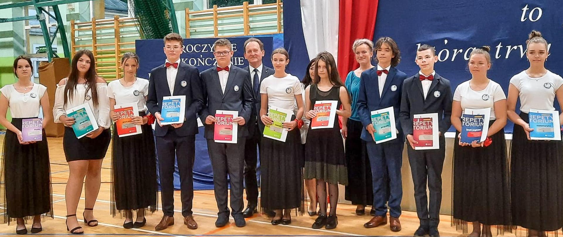 Na sali gimnastycznej stoi grupa kilkunastu elegancko ubranych osób, trzymają książki, za nimi wiceminister Bernacki.