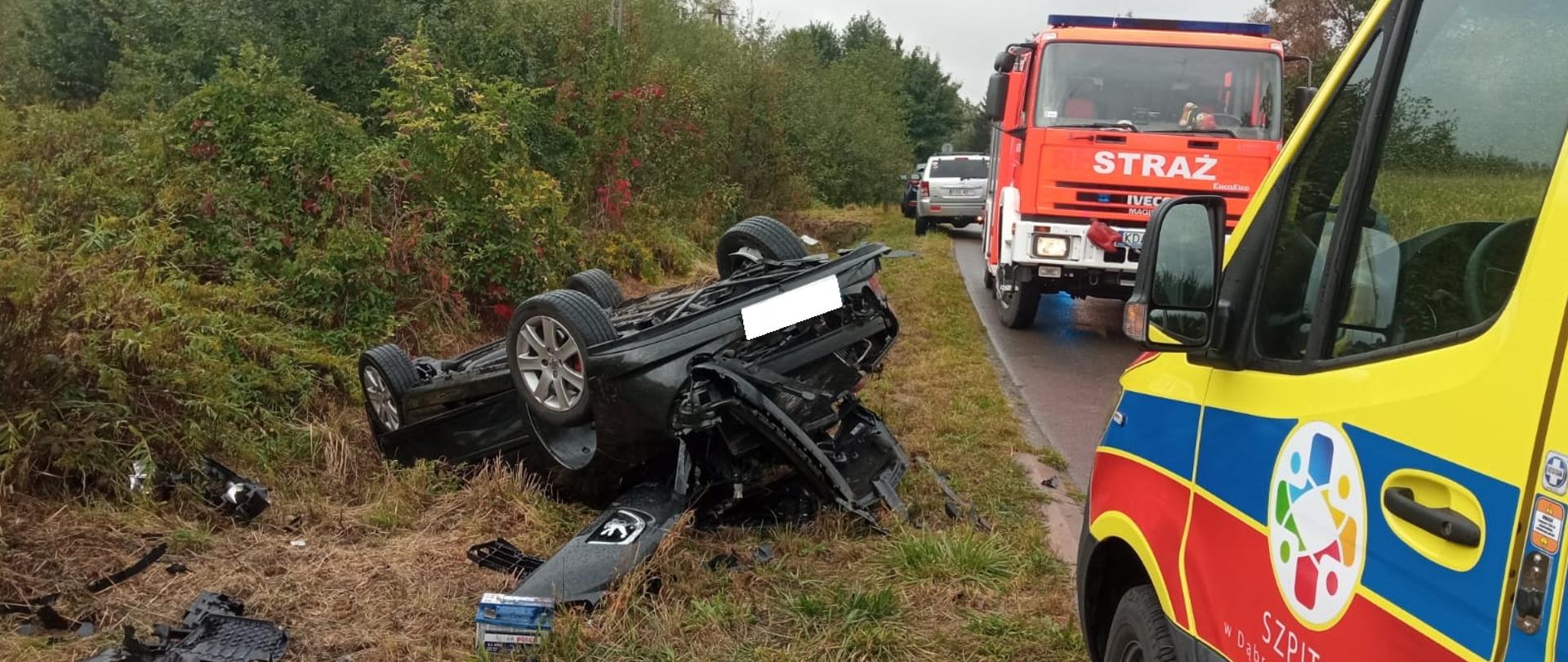 Na zdjęciu widać samochód osobowy po dachowaniu, obok pojazdy ratownicze.