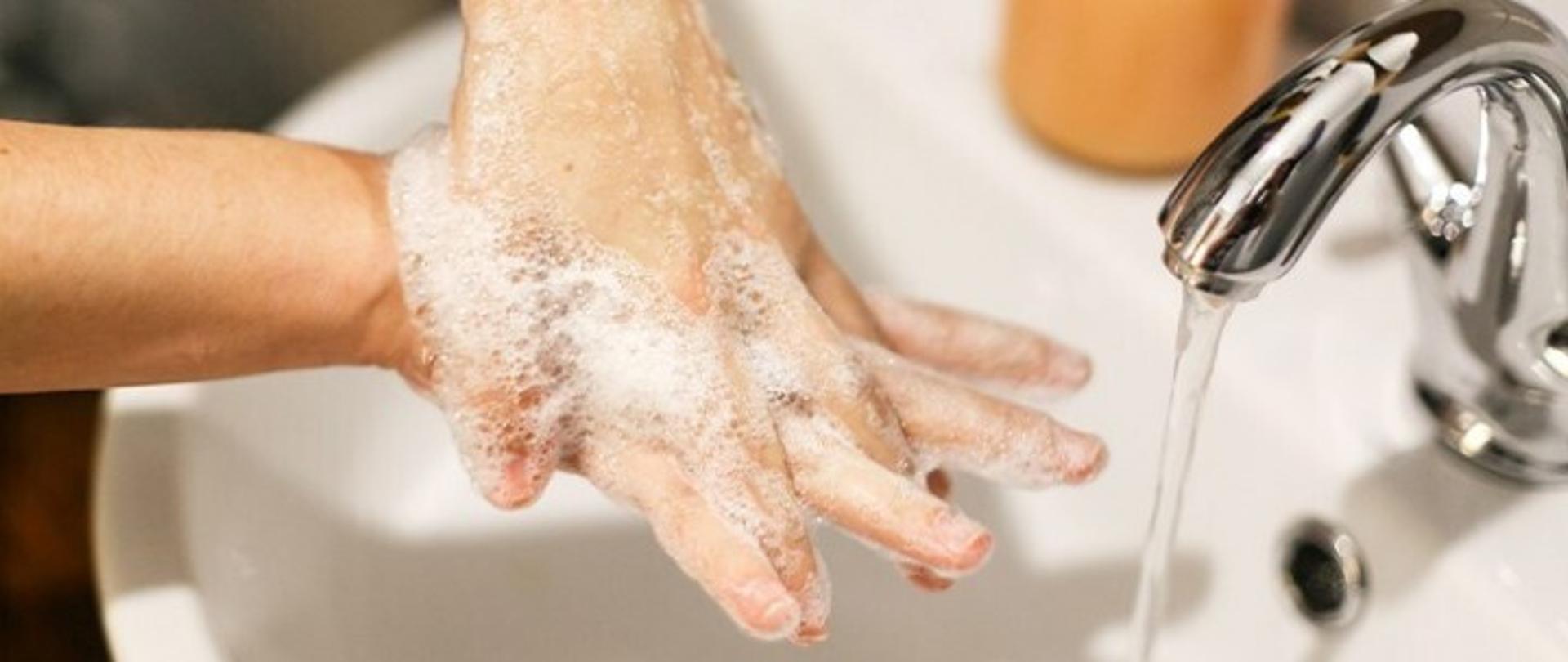 zdjęcie przestawiające mycie rąk, na głównym planie biała porcelanowa umywalka z srebrnym kranem, woda leci nieprzerwanym strumieniem. nad zlewem widoczne dłonie całe w pianie, jedna dłoń na drugiej, dłoń będąca na wierzchu myje przestrzenie między palcami dłoni pod nią.