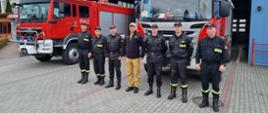 Zdjęcie przedstawia zastępcę komendanta głównego Państwowej Straży Pożarnej nadbrygadiera Arkadiusza Przybyłę, komendanta powiatowego PSP w Jędrzejowie mł. bryg. Mariusza Stolarczyka oraz zmianę służbową jednostki ratowniczo-gaśniczej. Wszyscy znajdują się przed bramami garażowymi na tle samochodów ratowniczo-gaśniczych.