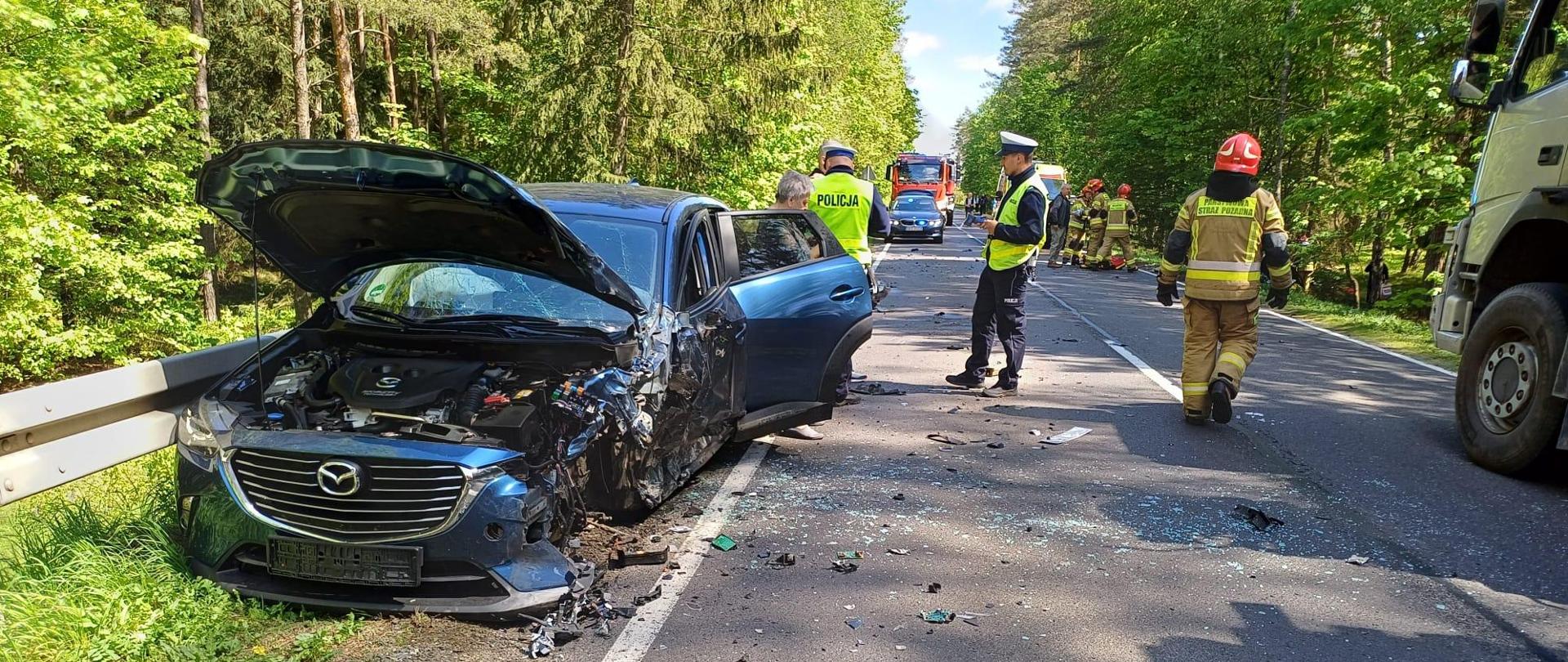 Zniszczony samochód na drodze z pracującymi służbami ratunkowymi.