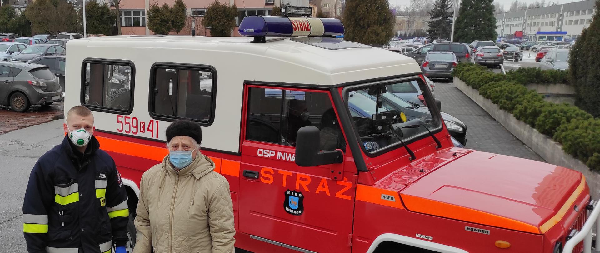 Po lewej stronie strażak w ubraniu specjalnym i maseczce na twarzy. Obok starsza kobieta wspierająca się laską z maseczką na twarzy w kurtce zimowej. Za nimi samochód terenowy samochód pożarniczy.