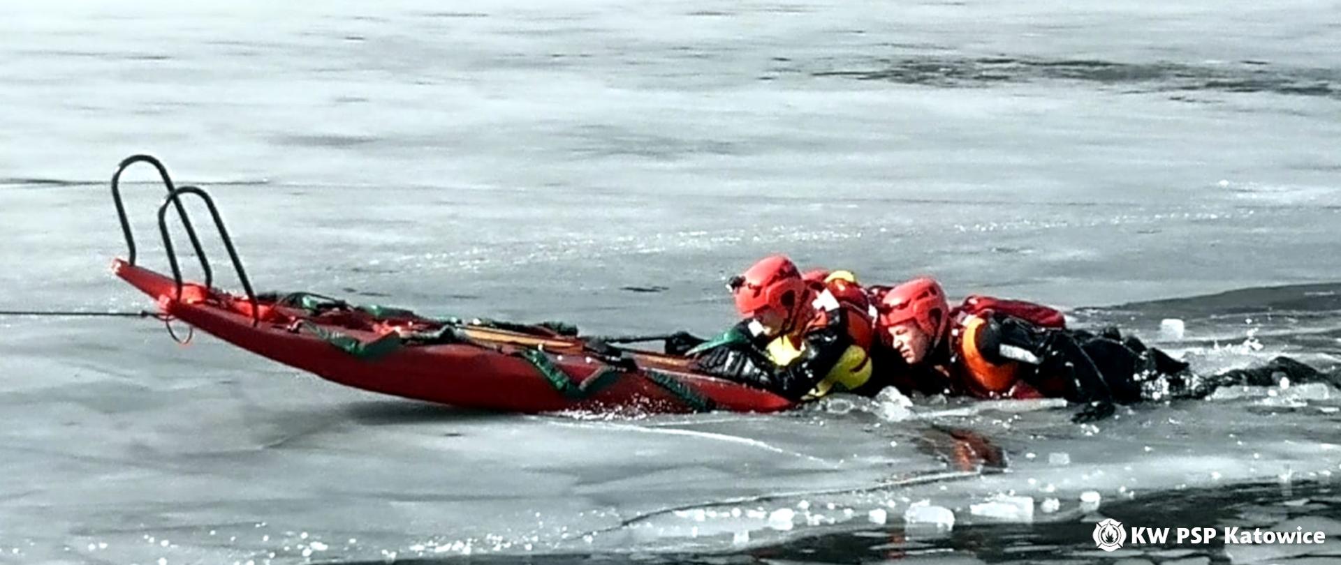 Na zdjęciu widoczny jest zamarznięty akwen i dwóch strażaków w skafandrach, którzy wciągają się z wody na sanie lodowe . 