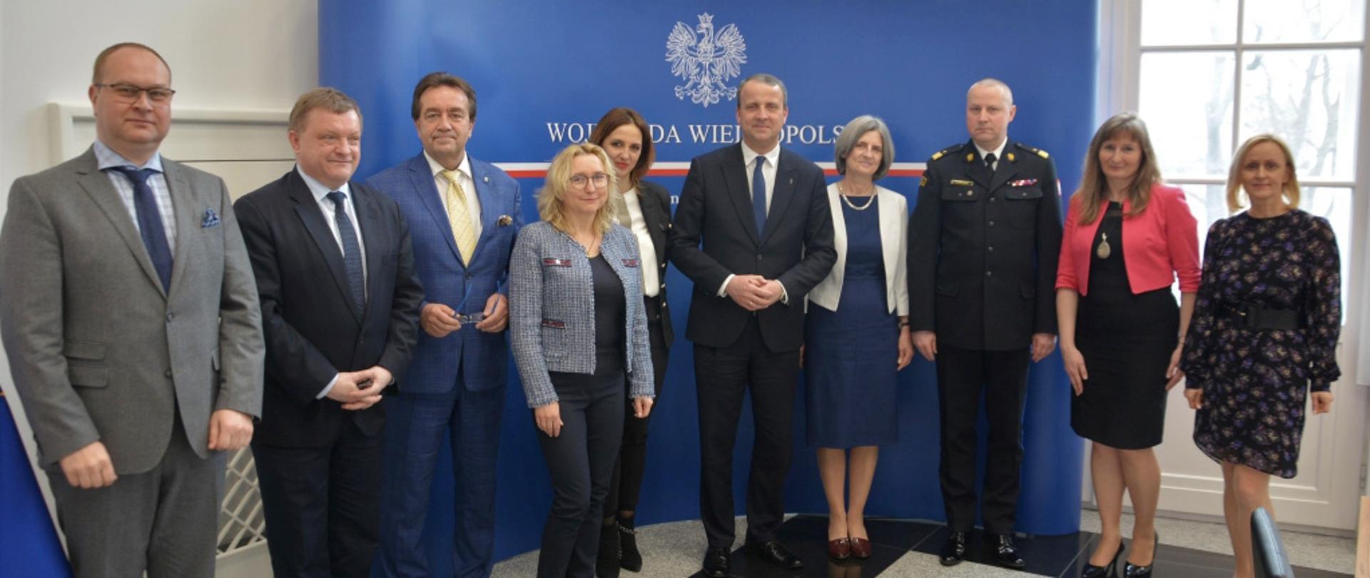 grupa ludzi stoi w szeregu , w tle niebieska ścianka reklamowa z orłem