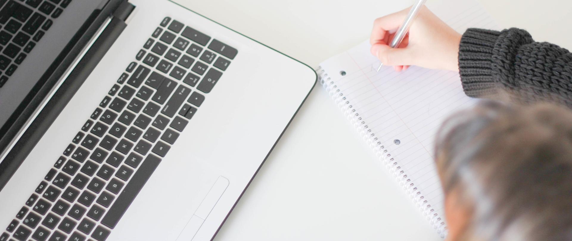 Srebrny laptop z czarnymi klawiszami na białym stole. W prawym dolnym rogu widoczna dziewczyna, która robi notatki w zeszycie w linie. 