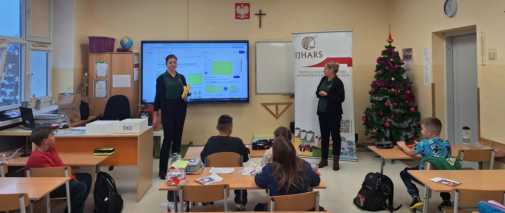 Inspektorki IJHARS prowadzą lekcje ekologii. W tle logo IJHARS i tablica multimedialna, w ławkach siedzą uczniowie.