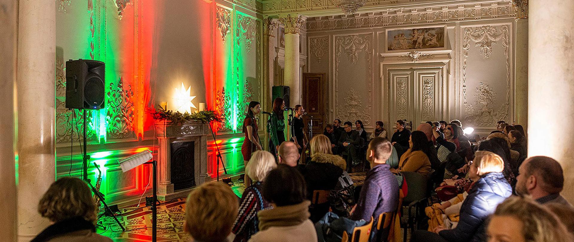 Zdjęcie przedstawiające uczniów PLSP w Kościelcu śpiewających w balowej sali szkoły oraz publiczność. Za uczniami ściana rozświetlona w czerwono zielonych barwach.