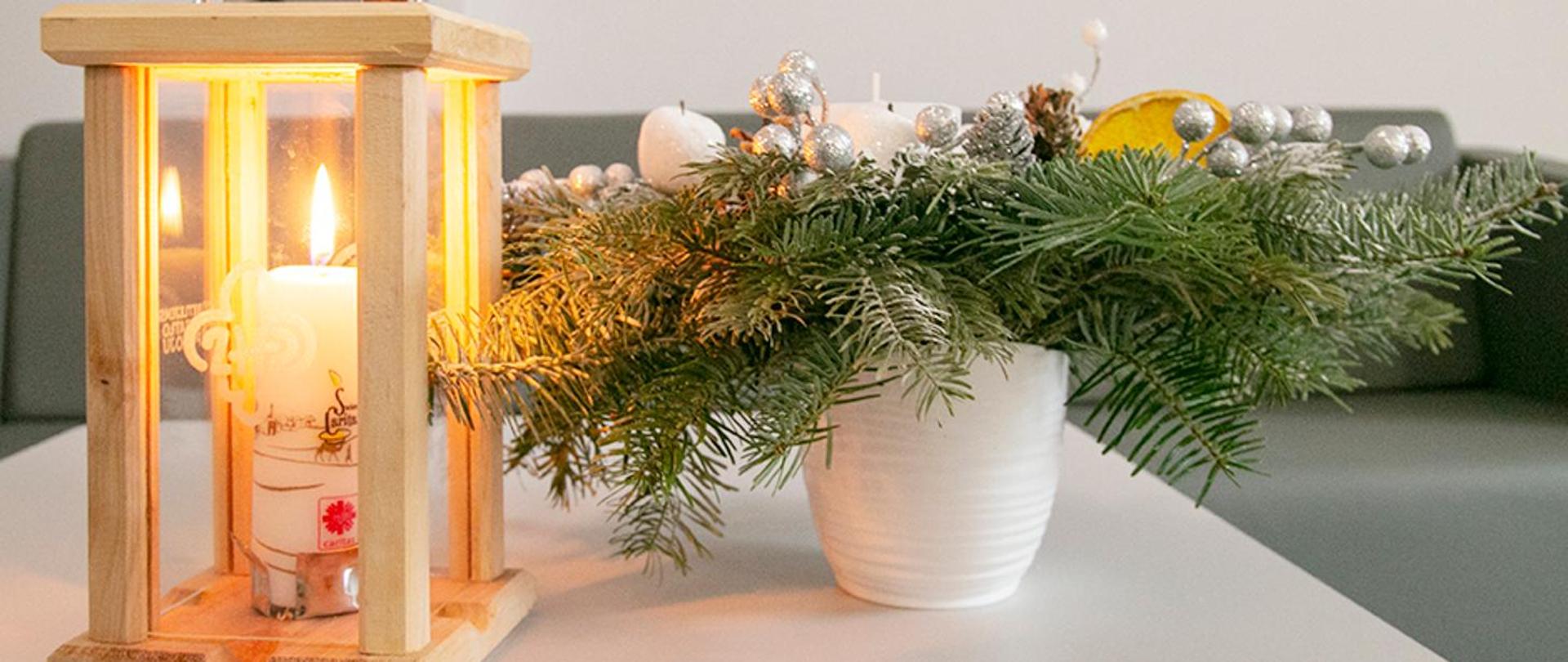Zdjęcie przedstawia stroik z gałązek świerkowych i drewniany lampion ze świecą. 