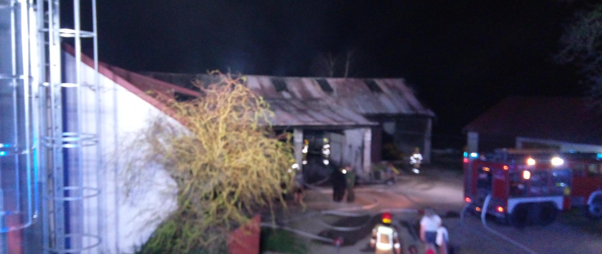 Budynek gospodarczo inwentarski w tle. Przed budynkiem samochód gaśniczy, strażacy oraz właściciele budynku. 