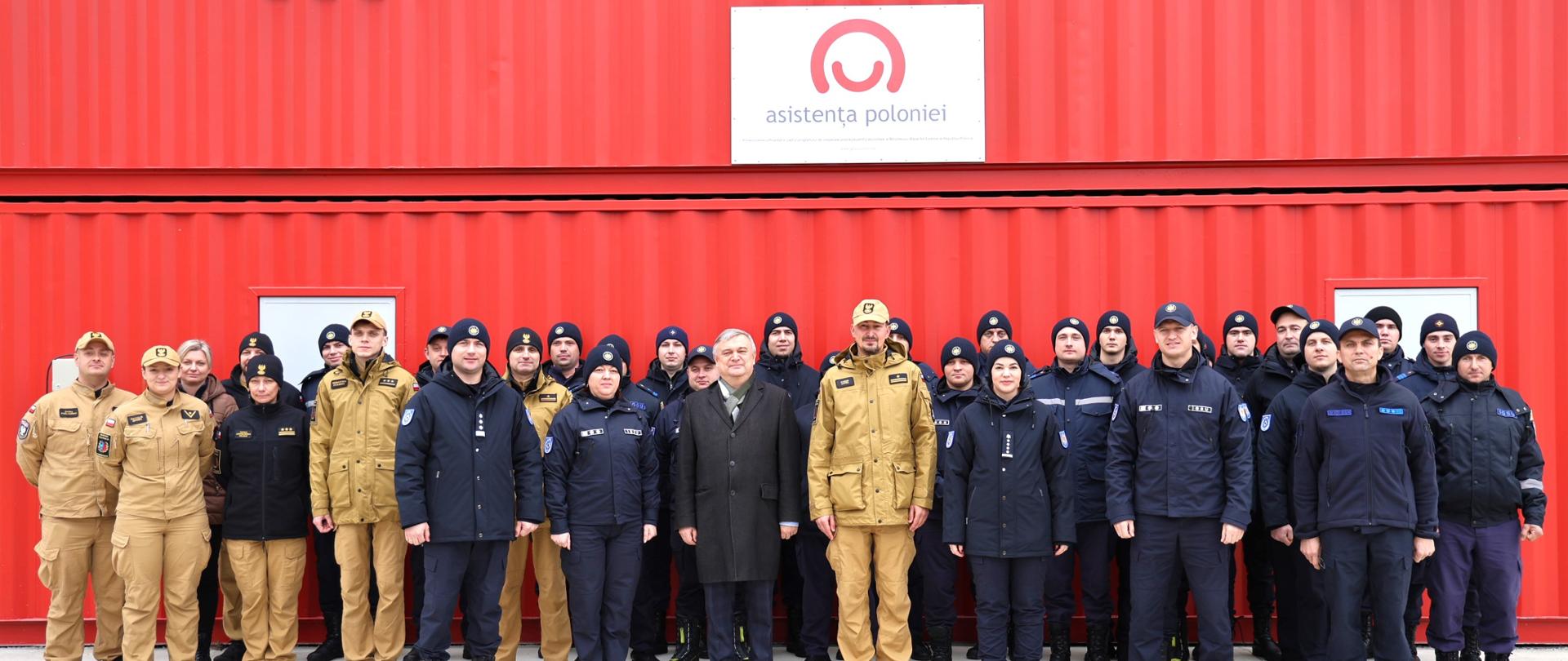 Grupowe zdjęcie uczestników szkolenia w służbowych mundurach na tle czerwonego kontenera z logo Polskiej pomocy po rumuńsku