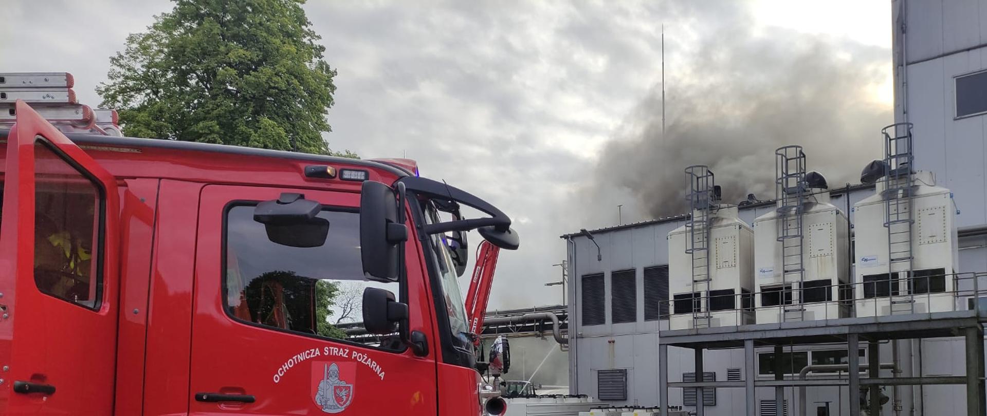 Na zdjęciu po lewej stronie widać samochód pożarniczy, po prawej w oddali widać budynek produkcyjny z którego z dachu wydobywa się dym.