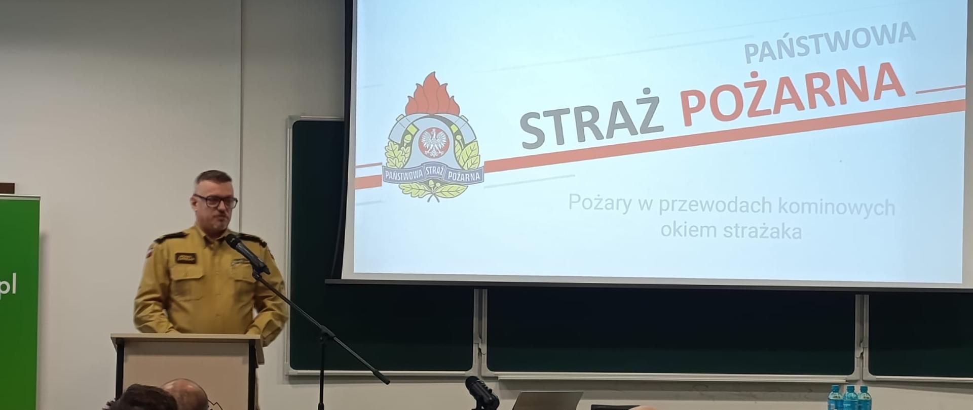 W jasnej sali wykładowej stoi przy mównicy st. bryg. Michał Zawiślak w beżowym mundurze Państwowej Straży Pożarnej. Mówi do mikrofonu, prezentując statystyki dotyczące pożarów w przewodach kominowych podczas IX Konferencji „Bezpieczne Ciepło 2026”. Za nim widoczny jest duży ekran z logotypem Państwowej Straży Pożarnej oraz slajdem zatytułowanym „Pożary w przewodach kominowych okiem strażaka”. W pierwszych rzędach siedzą uczestnicy konferencji — strażacy z województwa opolskiego zajmujący się prewencją pożarową