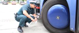 Inspektor mazowieckiej Inspekcji Transportu Drogowego kontroluje autokar turystyczny przed wyjazdem dzieci i młodzieży na wakacyjny wypoczynek. Funkcjonariusz sprawdza ogumienie i układ kierowniczy pojazdu.